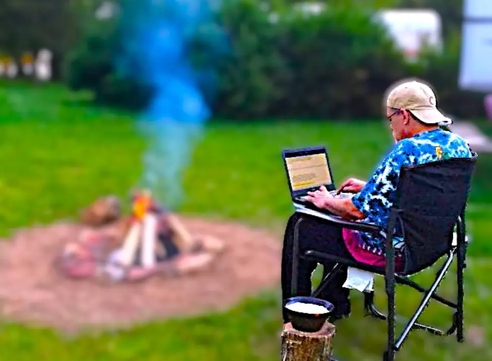 Man using laptop by campfire outdoors.